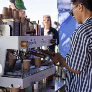 A barista wearing sunglasses and a striped shirt pulls espresso shots from a white outdoor coffee machine with wooden handles, with dozens of paper cups lined up on the machine and festival-goers milling in the background. Location: Taranaki, New Zealand